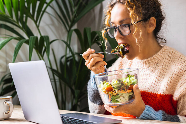 Jonge werknemer die salades eet in een snelle lunchpauze terwijl hij zonder onderbreking aan de laptop werkt. Drukke zakenvrouw die gezond eet en laptop op tafel gebruikt. Kantoor en bedrijf. Gezondheid levensstijl baan - Foto, afbeelding