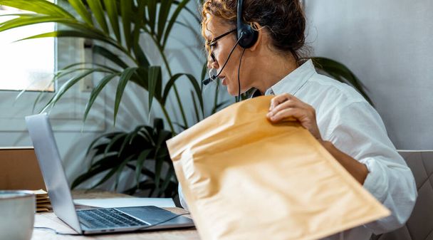 Employee female working at the desk talking with clients using headset and microphone and laptop. Customer care service business. One adult woman work online and prepare package for shipment remote - Φωτογραφία, εικόνα