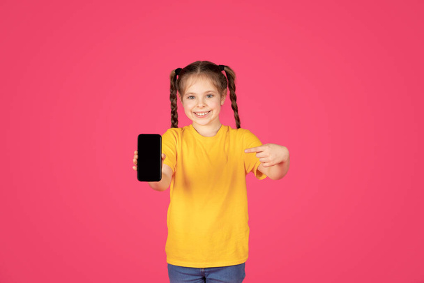Cute Preteen Girl Pointing At Smartphone With Blank Screen In Hand, Cheerful Female Child Advertising New Mobile Application Or Website, Standing Isolated Over Pink Background, Mockup Image - Zdjęcie, obraz