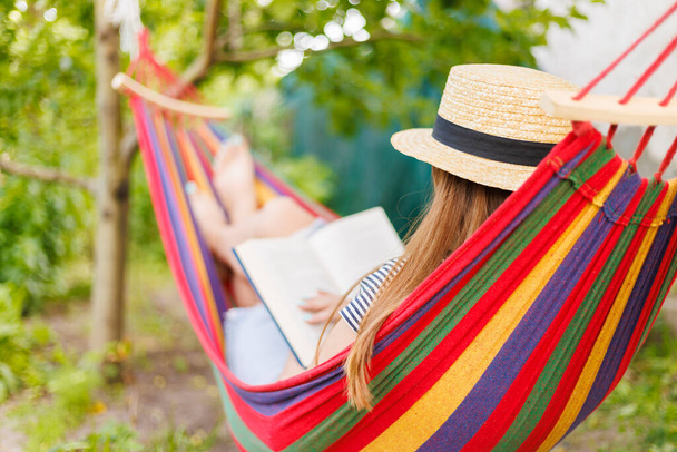 Young woman reading book while lying in comfortable hammock at green garden. - Photo, Image
