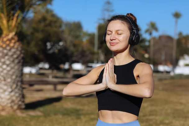 Young healthy woman in sportswear listens music in headphones while doing fitness squat leg exercises outdoors in park with palm trees. Workout for strengthen pelvic floor muscles on summer holiday. - Photo, Image