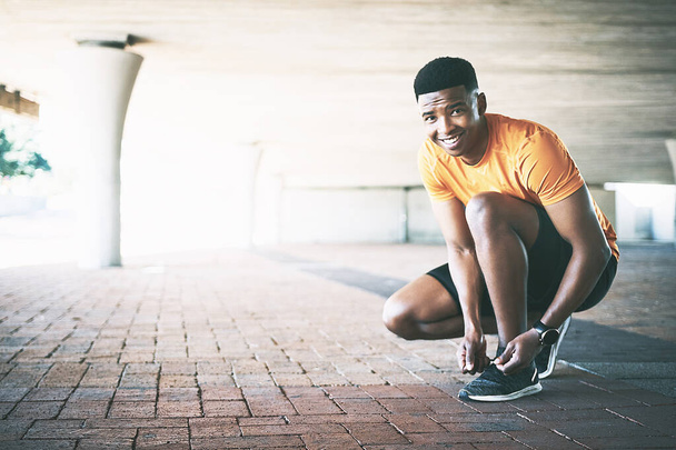 Aan de slag voor een fitter, gezonder, gelukkiger jij. Portret van een jongeman die zijn veters vastbindt tijdens een training tegen een stedelijke achtergrond - Foto, afbeelding