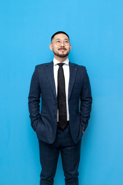 komoly sikeres ázsiai üzletember öltöny és szemüveg áll kék elszigetelt háttér és felnéz, koreai vállalkozó üzleti ruhák néz a másolás tér, függőleges keret - Fotó, kép