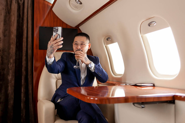 asiatico uomo d'affari in giacca e cravatta e occhiali con bicchiere di champagne vola in jet di lusso e utilizza smartphone, coreano imprenditore si filma al telefono e comunica tramite videochiamata - Foto, immagini