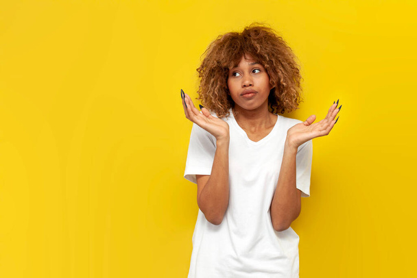 νεαρό αβέβαιο σγουρό αμερικανικό κορίτσι shrugging τους ώμους της σε κίτρινο απομονωμένο φόντο, Αφρικανή γυναίκα σε λευκό t-shirt δεν ξέρει και απλώνει τα χέρια της στα πλάγια - Φωτογραφία, εικόνα