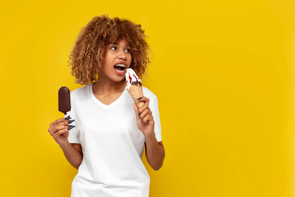 young curly american girl with braces holding two different ice creams with jam and daydreaming over yellow isolated background, african woman with toothy smile chooses chocolate or sundae - 写真・画像