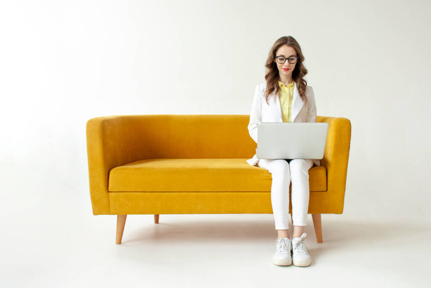 若いビジネスマンスーツの女性は快適な柔らかいソファに座って、ラップトップを使用して、正式な摩耗の女の子は白い隔離された背景に黄色のソファの上にコンピュータに入力されています - 写真・画像