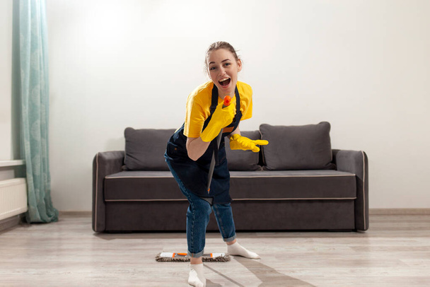 νεαρή ευτυχισμένη νοικοκυρά κορίτσι με ποδιά και γάντια πλένει το πάτωμα με σφουγγαρίστρα και χορούς, γυναίκα ακούει μουσική και καθαρίζει το δωμάτιο, οικονόμος με στολή τραγουδά και κάνει υγρό καθαρισμό - Φωτογραφία, εικόνα