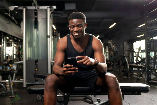 young athletic african american man sits in dark gym and uses smartphone, sports guy types online on the phone in fitness club and smiles - Foto, immagini