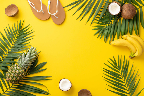 Embrace the summer holiday vibe. Top view showcasing beach gear, flip-flops, palm leaves, and juicy exotic fruits against a vivid yellow background with frame for text or advertisements - Fotografie, Obrázek