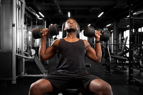 若い運動アフリカ系アメリカ人男性がダークジムでトレーニングし、運動選手はフィットネスクラブで重いダンベルを持ち上げる - 写真・画像