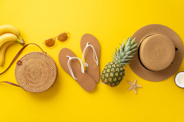 Embracing the summer's arrival. Aerial top view shot of beach gear, sunglasses, sunhat, sandals, rattan handbag, starfish, juicy pineapple, coconut and bananas on a lively yellow background - Photo, Image