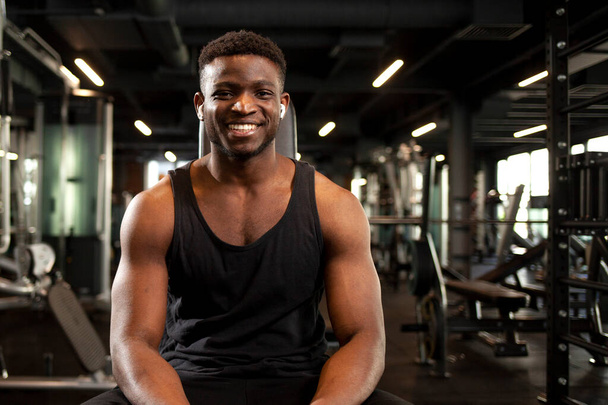 young athletic african american man sits in dark gym and smiles, athletic guy rests and looks at the camera in fitness club, portrait of athlete in training - Fotó, kép