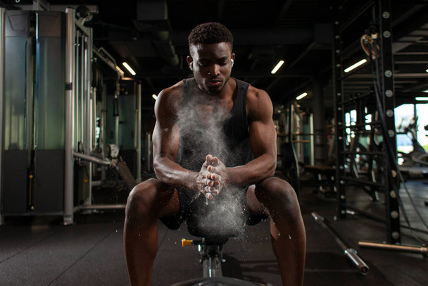 sportovní afroameričan muž vlaky v tmavé tělocvičně a tře si ruce s magnézií, mladý chlap dělá tleskat s rukama v posilovně, detailní záběr prachu a mouky s rukama atleta - Fotografie, Obrázek