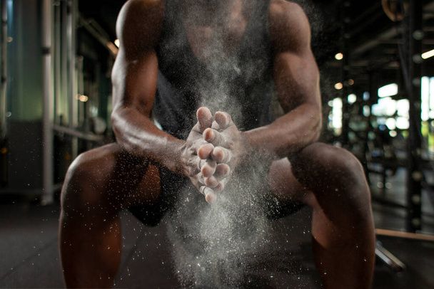 sportlicher afrikanisch-amerikanischer Mann trainiert in dunkler Turnhalle und reibt sich die Hände mit Magnesia, junger Kerl klatscht mit den Händen im Fitnessraum, Nahaufnahme von Staub und Mehl mit den Händen des Athleten - Foto, Bild