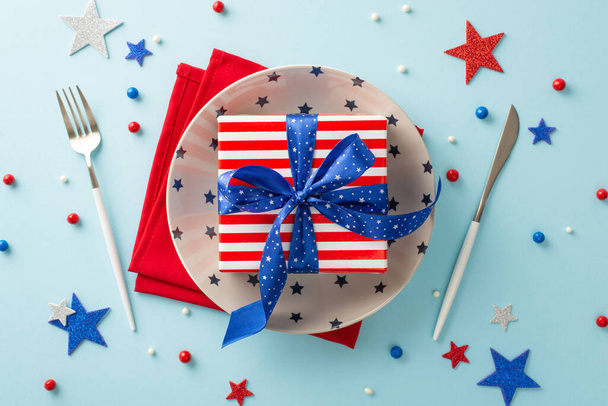 ¡Haz brillar tu mesa en el cumpleaños de Estados Unidos! Vista superior de conjunto patriótico con caja de regalo en el envoltorio con patrón de bandera de EE.UU. en la placa, cubiertos, servilleta, aspersiones, estrellas sobre un fondo azul pastel - Foto, Imagen