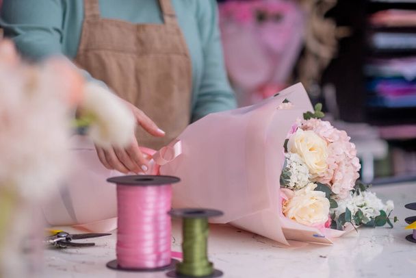 At the florists, a young woman in her apron, skillfully putting together beautiful flowers and green branches mixing bouquets and carefully wrapping them in Kraft paper. - Foto, Imagem