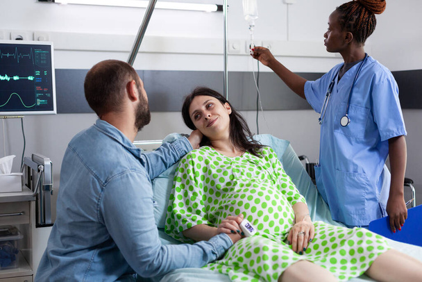 Ζευγάρι με εγκυμοσύνη περιμένει να γεννήσει στο νοσοκομείο. Αφροαμερικανή νοσοκόμα ελέγχει IV ορό τσάντα ενστάλαξης, ενώ έγκυος γυναίκα που βρίσκεται στο κρεβάτι και ο σύζυγος παρηγορεί για τη μητρότητα - Φωτογραφία, εικόνα