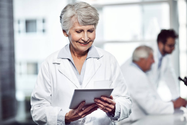 Echando un vistazo a las estadísticas. una científica anciana alegre usando una tableta digital mientras está de pie dentro de un laboratorio - Foto, Imagen