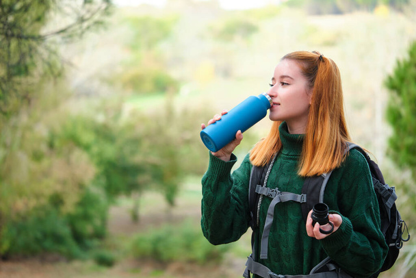 Smiley hiker redhead young woman drinking water from bottle in the mountain wearing a backpack. Trekking concept. - Photo, Image