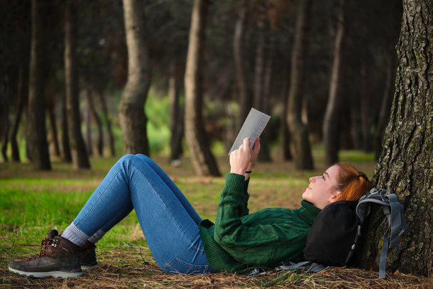 Redhead young woman reading a book lying under tree in forest. Relaxing in nature. - Valokuva, kuva
