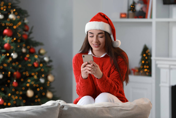 Smiling woman wearing red Christmas hat with smartphone in cosy room - Foto, imagen