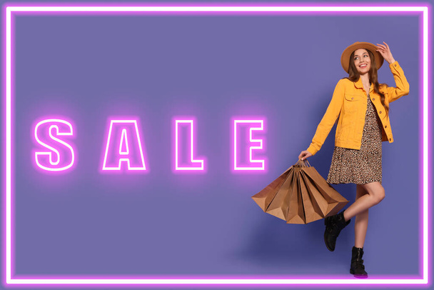 Stylische junge Frau mit Einkaufstaschen, Neon-Rahmen und Word Sale auf violettem Hintergrund - Foto, Bild