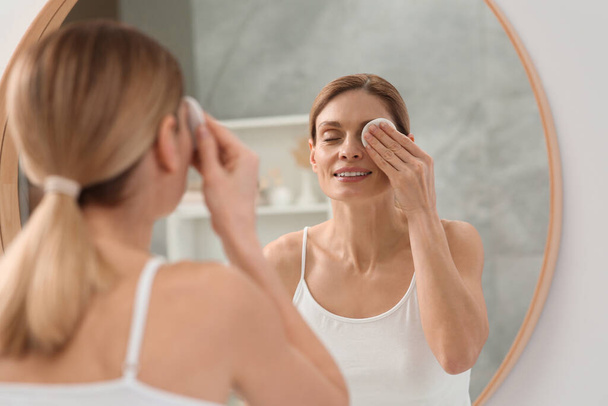 Beautiful woman removing makeup with cotton pad near mirror indoors - Foto, Imagem