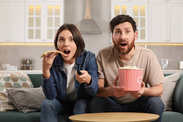 Surprised couple watching TV with popcorn and pizza on sofa at home - Φωτογραφία, εικόνα