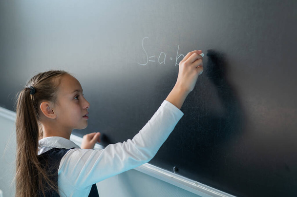 The schoolgirl answers at the lesson. Caucasian girl writes a formula on a blackboard - Фото, зображення