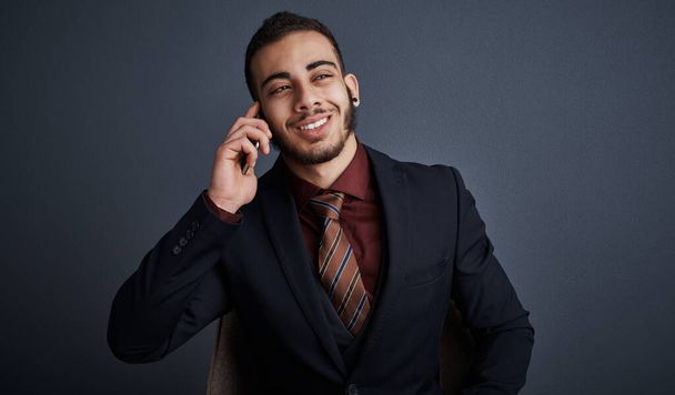 Give me the good news. Studio shot of a stylish young businessman looking thoughtful while making a phonecall against a gray background - Foto, Imagen