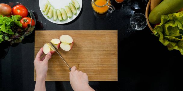 Couteau à main serrée coupant une pomme rouge sur une planche à découper en bois (vue du dessus). Les fruits tranchés sont placés sur une assiette. Le comptoir de cuisine plein de divers types de légumes et de jus de fruits. - Photo, image