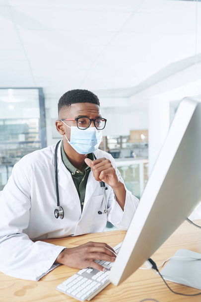 Ich bin ein wenig besorgt über diese Daten. ein junger Arzt mit Maske sitzt in seinem Büro in der Klinik, während er einen Computer benutzt - Foto, Bild
