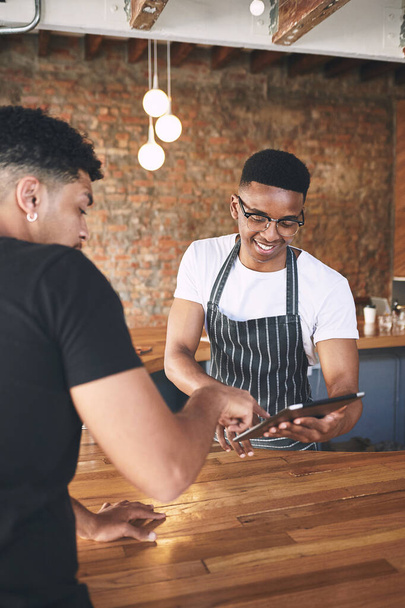 Ένας καφές έρχεται αμέσως. ένας νεαρός άνδρας που χρησιμοποιεί ένα ψηφιακό tablet ενώ σερβίρει έναν πελάτη σε ένα καφέ - Φωτογραφία, εικόνα