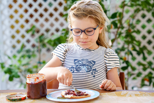 Cute funny preschool girl eats sweet bun for breakfast. Happy child eating bread roll with strawberry, cherry or plum jam. Health food for children and kids with selfmade jelly. - Photo, image