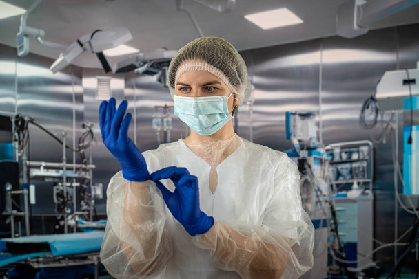 Una enfermera usa un uniforme azul con una máscara mientras se pone guantes azules de látex de goma durante una operación quirúrgica compleja. medicina. médicos quirófano - Foto, imagen