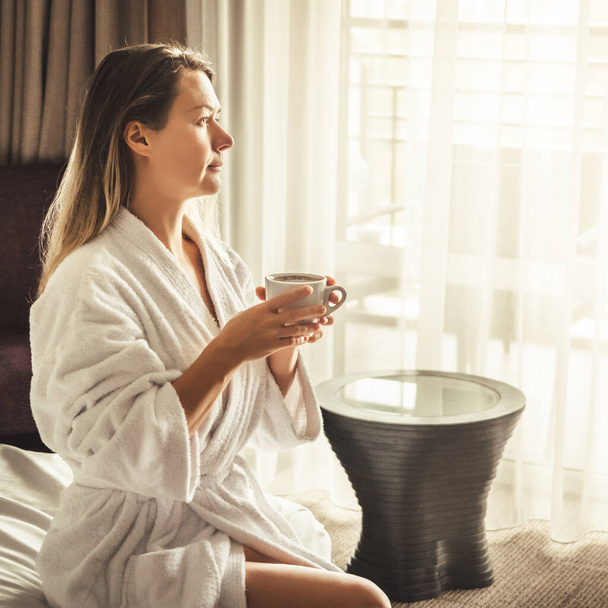 Woman in white bathrobe lying on sofa and relaxing with orange cocktail at the roof of hotel - Photo, Image