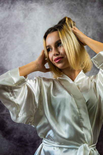 Hübsche junge Frau in heller Seidenrobe auf schlichtem Hintergrund posiert im Atelier. isolierte Heimkleidung Pyjama - Foto, Bild