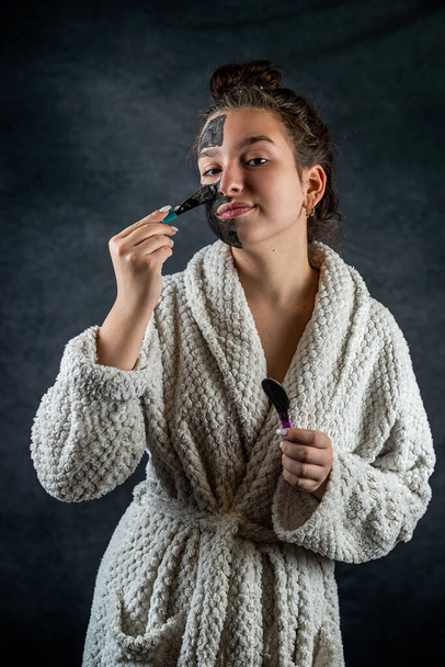 Young beautiful woman applies a restorative black mask with brush to her face isolated on dark. Health care lifestyle - Photo, image