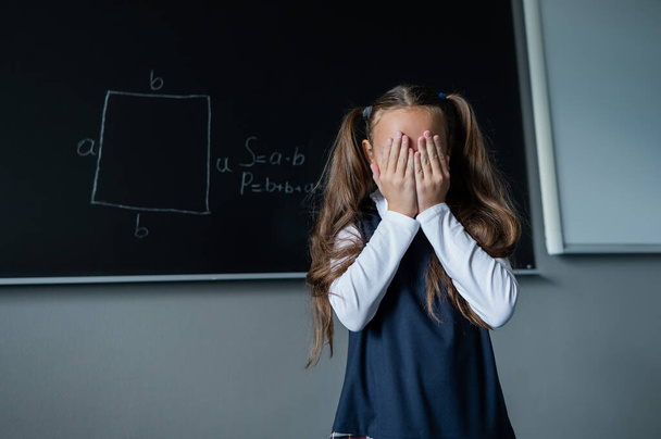 Caucasian girl stands at the blackboard and cries covering her face with her hands. The schoolgirl did not prepare for the lesson - 写真・画像
