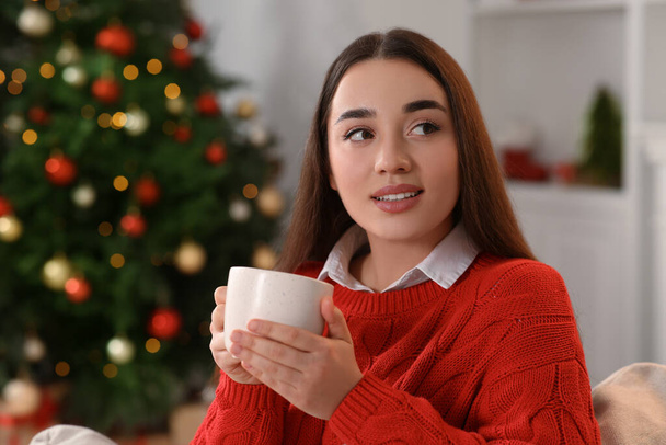 Woman holding cup of hot drink on sofa near Christmas tree indoors - 写真・画像
