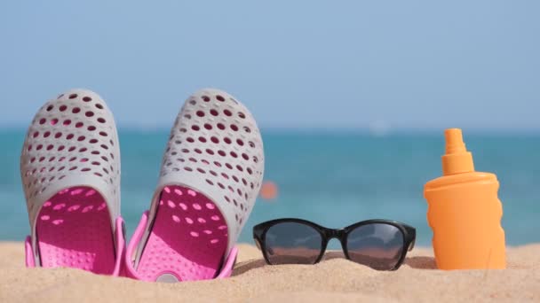 Closeup of clogs shoes, sunscreen and black protective sunglasses on sandy beach at tropical seaside on warm sunny day. Summer vacation concept. - Footage, Video