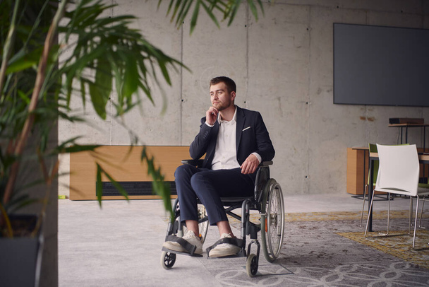 A melancholic businessman in a wheelchair sitting with a sad expression, gazing through the window of a modern office, conveying a sense of solitude.  - Foto, Imagen