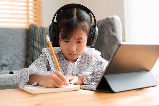Asian little girl doing homework, using digital tablet with headphone. Homeschooling, Concept online learning at home - Valokuva, kuva