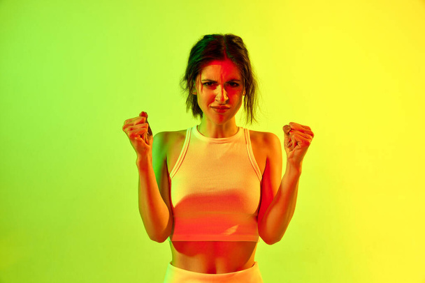 Portrait d'une charmante fille agacée montrant des poings avec un visage en colère sur fond de studio de couleur vert acide au néon. Concept de défis, émotions humaines, force, arts martiaux, publicité - Photo, image