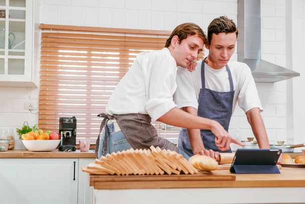 Heureux couple gay caucasien cuisiner ensemble dans la cuisine tout en regardant comment cuisiner sur une tablette numérique avec plaisir et un sourire. Un homme prêt à préparer à manger à la maison. Concept de couple LGBT. - Photo, image
