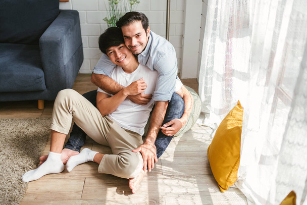 Белая и азиатская ЛГБТ-пара обнимаются сзади, сидят вместе дома. Разнообразие ЛГБТ-отношений. Концепция совместной жизни гей-пары. - Фото, изображение