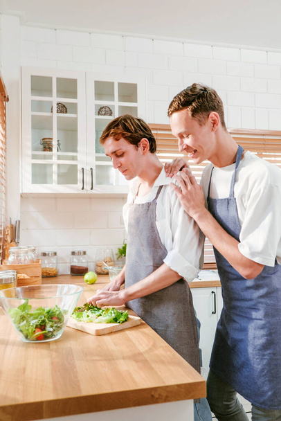 Glückliches kaukasisches schwules Paar, das gemeinsam Salat zubereitet Eine Person hat mit einem Messer Salat davor geschnitten. Und eine andere Person, die zu Hause in der Küche von hinten zuschaut. LGBT-Beziehungen. Konzept für schwule Paare - Foto, Bild