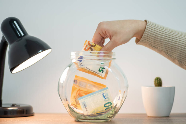 Weibliche Hand wirft Geld in ein Glas für eine Ersparnis. Euro-Banknoten als Investition in die Zukunft, persönliches und familiäres Wirtschaftskonzept - Foto, Bild