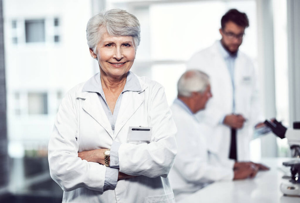 Она уже некоторое время работает в медицинской индустрии. Портрет веселой пожилой женщины-ученого, стоящей со сложенными руками и смотрящей в камеру внутри лаборатории - Фото, изображение